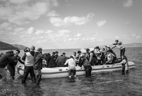 Lesbos, Greece - October 25, 2015: Volunteer lifeguards and others assist migrants out of their boat after landing on the Greek island of Lesbos, near the town of Skala Sikamineas. A photographer (left) photgraphs the landing. The coastline of Turkey is visible on the rightside of the photo.
