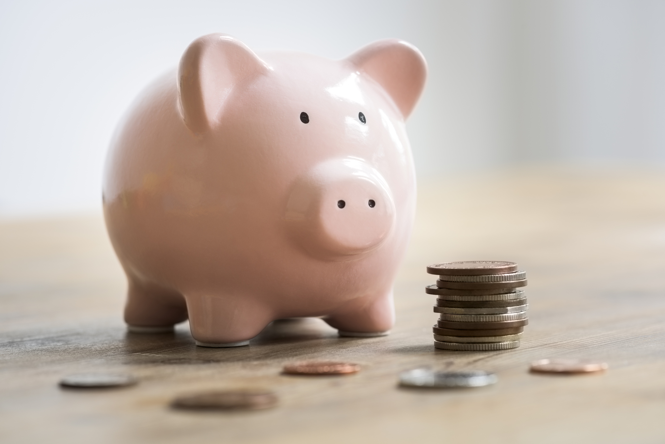 Loose coins and a stack of coins are lying on a wooden table in front of a pink ceramic piggy bank. The selective focus is on the piggy banks face. The coins are bronze and silver and are to be deposited into the piggy bank as savings.