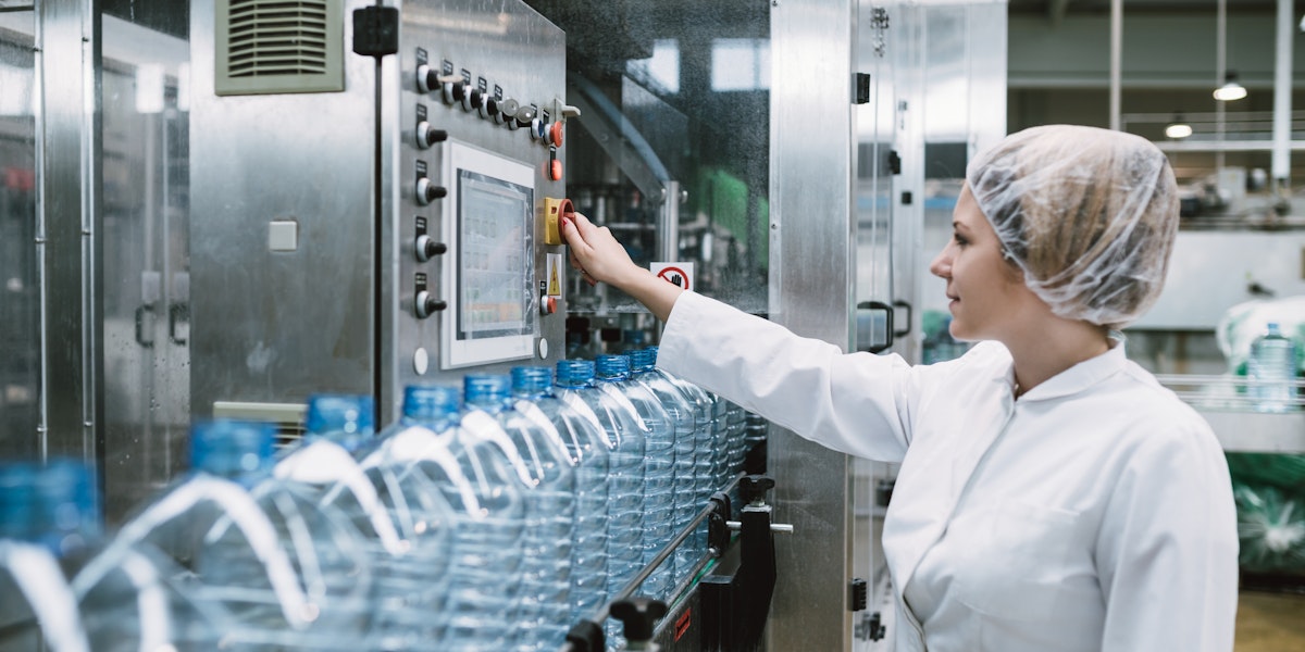 Young happy woman worker checking robotic line for bottling and packaging pure drinking water into bottles and canisters.