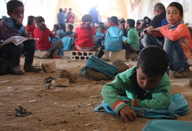On 13 November 2016, children lie or sit on the ground writing in notebooks at a make-shift school in rural Dar'a in the Syrian Arab Republic. Despite the ongoing violence across the country, children and dedicated teachers are doing all they can to keep their education going. Some 1.7 million children are currently (Nov 2016) out of school in the Syrian Arab Republic. Schools across the Syrian Arab Republic continue to be destroyed and damaged. This year (2016) 84 attacks on schools have resulted in the deaths of 69 children. UNICEF has provided 3.2 million children with learning materials, such as school bags, stationary and text books.

In November 2016, there is no safe place for children to learn or play in Syria. Underground facilities including basements and even caves, are used to shelter children from a war they grew up knowing nothing else. Displaced families from different areas in rural Damascus and Dar’a sought refuge in tents in rural Dar’a. The make-shift school receives up to 80 children on a daily basis. They are grouped according to age and knowledge in four groups, where each of the four former teachers, displaced themselves, teaches them reading and writing in both Arabic and English and the principals of mathematics. “There is not a single space or even an extra tent that we could’ve used as a learning space,” says [NAME CHANGED] Muhammad, a teacher in this school. “So we had to clean this fodder collection centre and turn it to a school where almost 80 children come on a daily basis to learn reading and writing, both in Arabic and English, and the principals of mathematics,” Muhammad continues.  The children are divided into four groups based on their knowledge and age. Each group is taught by one of the four former teachers, two male and two female teachers. “Children have to share notebooks because we severely lack the necessary education supplies. They rotate on using the only six desks available.”  [NAME CHANGED] Mona, a