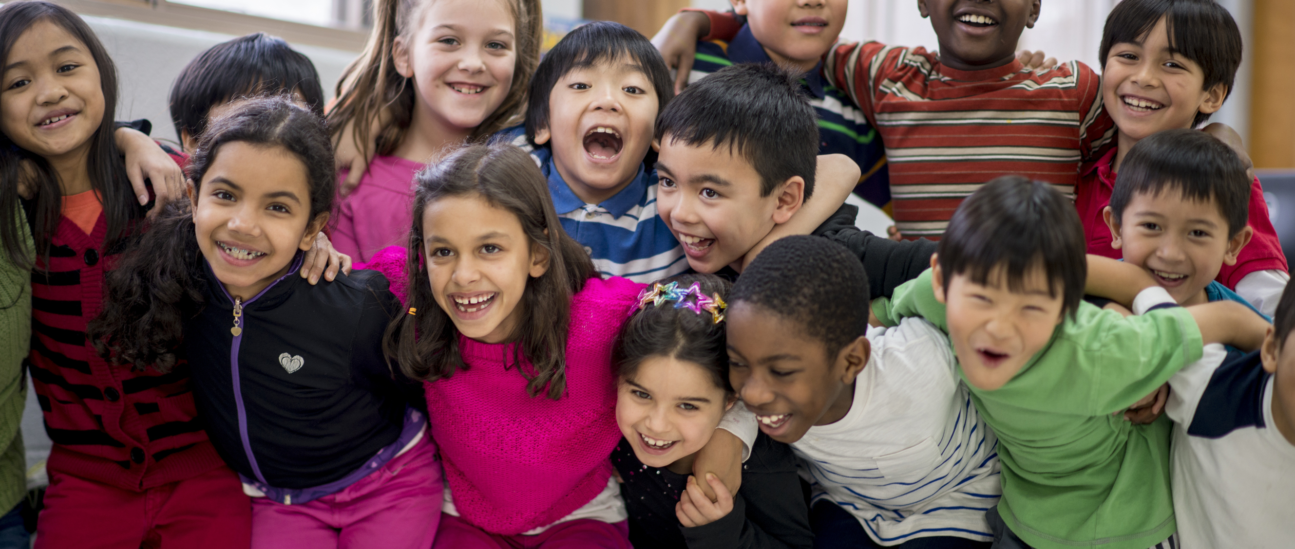 A multi-ethnic group of elementary school children are indoors in a classroom. They are wearing casual clothing. The students are standing and taking a fun group photo together.