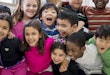 A multi-ethnic group of elementary school children are indoors in a classroom. They are wearing casual clothing. The students are standing and taking a fun group photo together.
