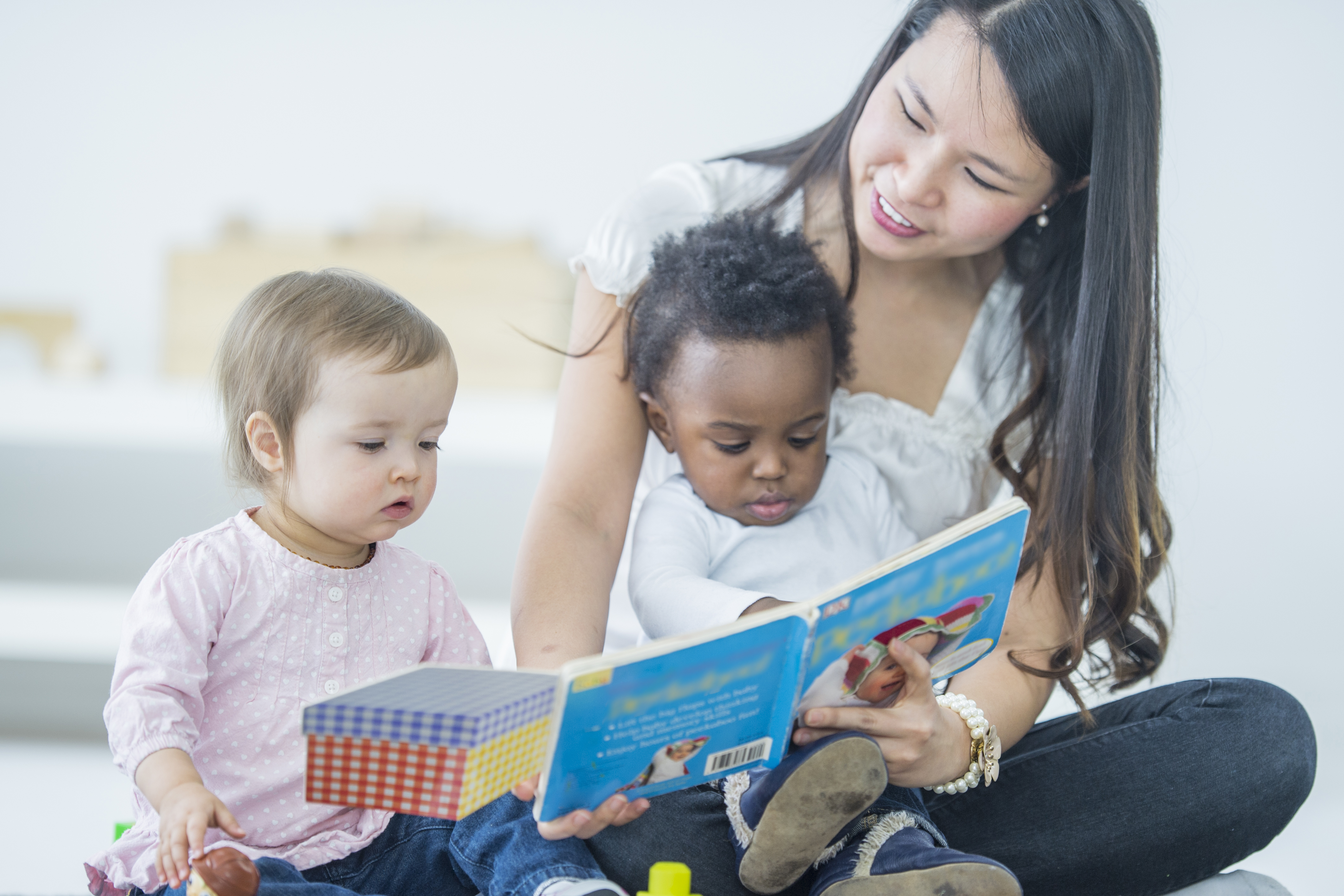 Young Asian woman reading to two babies. The woman is sitting on the floor focusing on the book and reading while holding one child in her lap and one child sitting on the carpet next to the woman. Two children sitting with care taker, one baby of caucasian ethnicity and one baby of African Descent