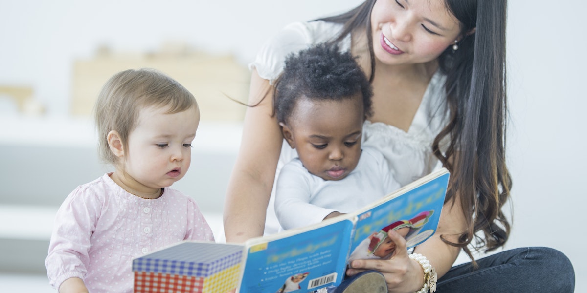Young Asian woman reading to two babies. The woman is sitting on the floor focusing on the book and reading while holding one child in her lap and one child sitting on the carpet next to the woman. Two children sitting with care taker, one baby of caucasian ethnicity and one baby of African Descent