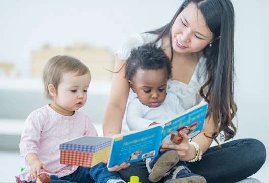 Young Asian woman reading to two babies. The woman is sitting on the floor focusing on the book and reading while holding one child in her lap and one child sitting on the carpet next to the woman. Two children sitting with care taker, one baby of caucasian ethnicity and one baby of African Descent