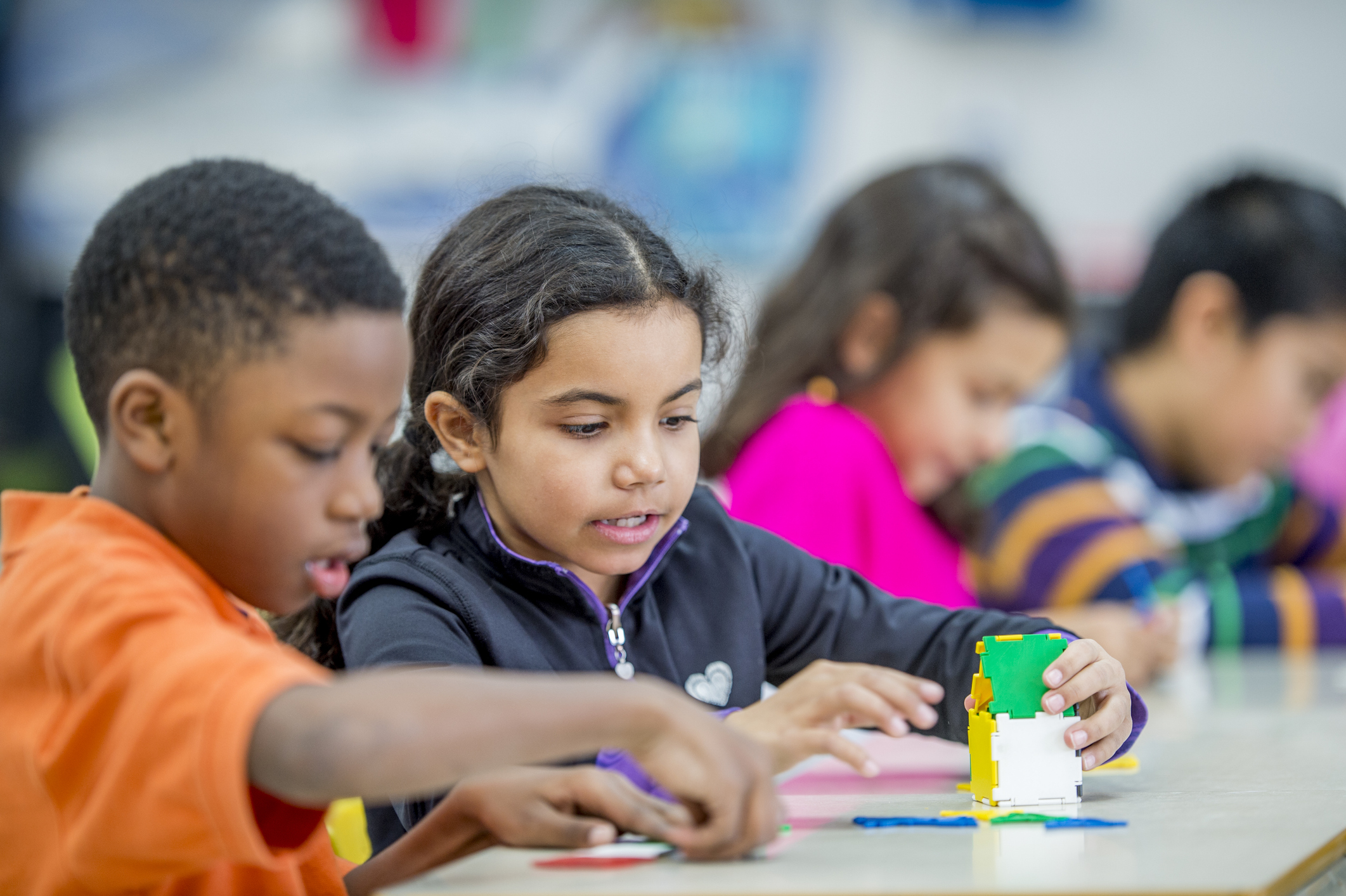 A multi-ethnic group of elementary school children are indoors in their classroom. A boy and girl are sitting at a table and playing with toy building tiles together.