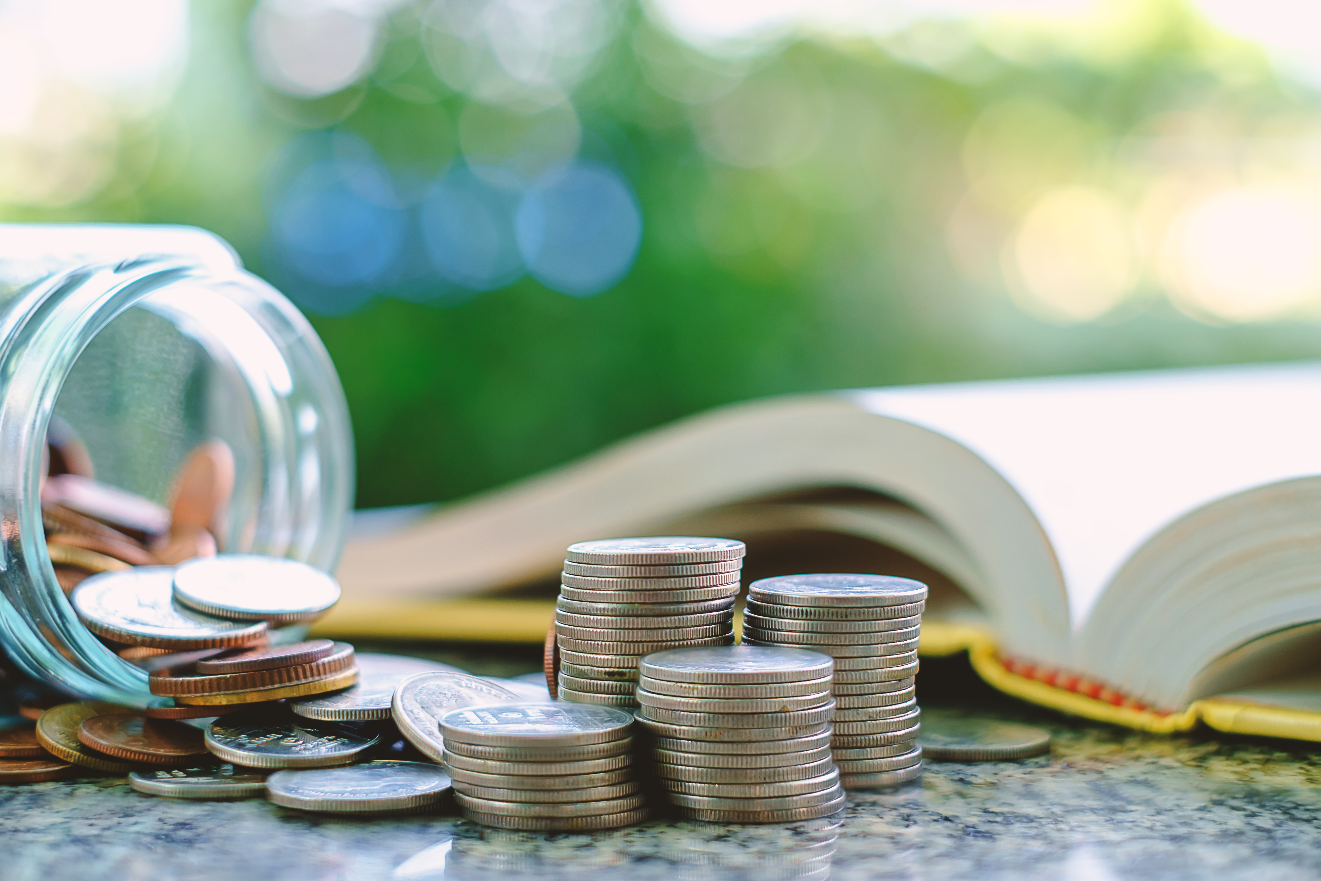 Pile of money coins in and outside the glass jar on blurred book and natural green background for financial and education concept