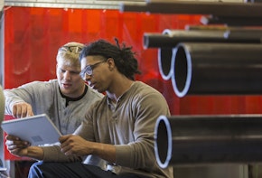 Two young multi-ethnic men using a digital tablet.  They are in a factory or workshop. The mixed race man is holding the tablet, and the other man is pointing to the screen.