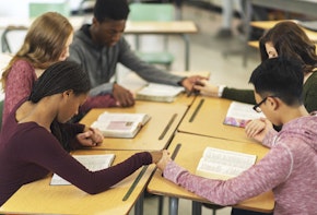 Five students in a classroom are sitting with their desks pushed together. They are holding hands in a circle and have their heads bowed in prayer. There are bibles open on their desks.