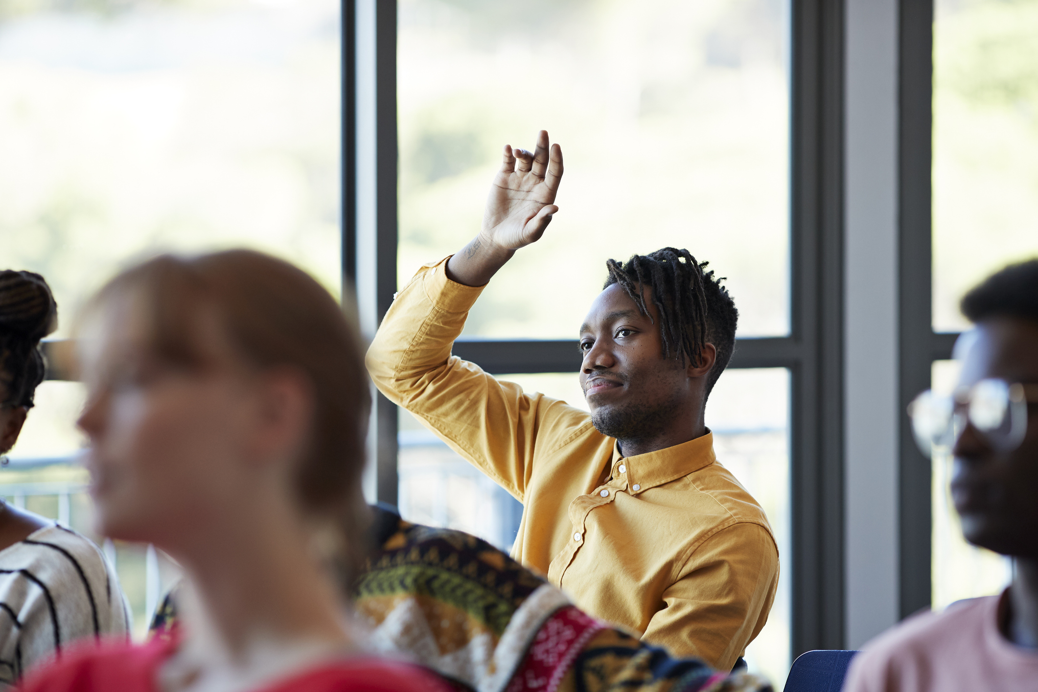 Confident young male student looking away while sitting with arm raised amidst friends in classroom against window at community college