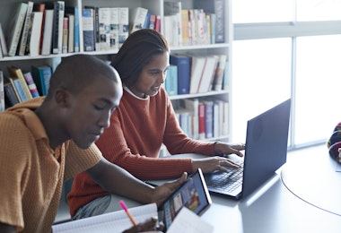 Young woman using laptop while sitting by friend studying at desk against bookshelf in library