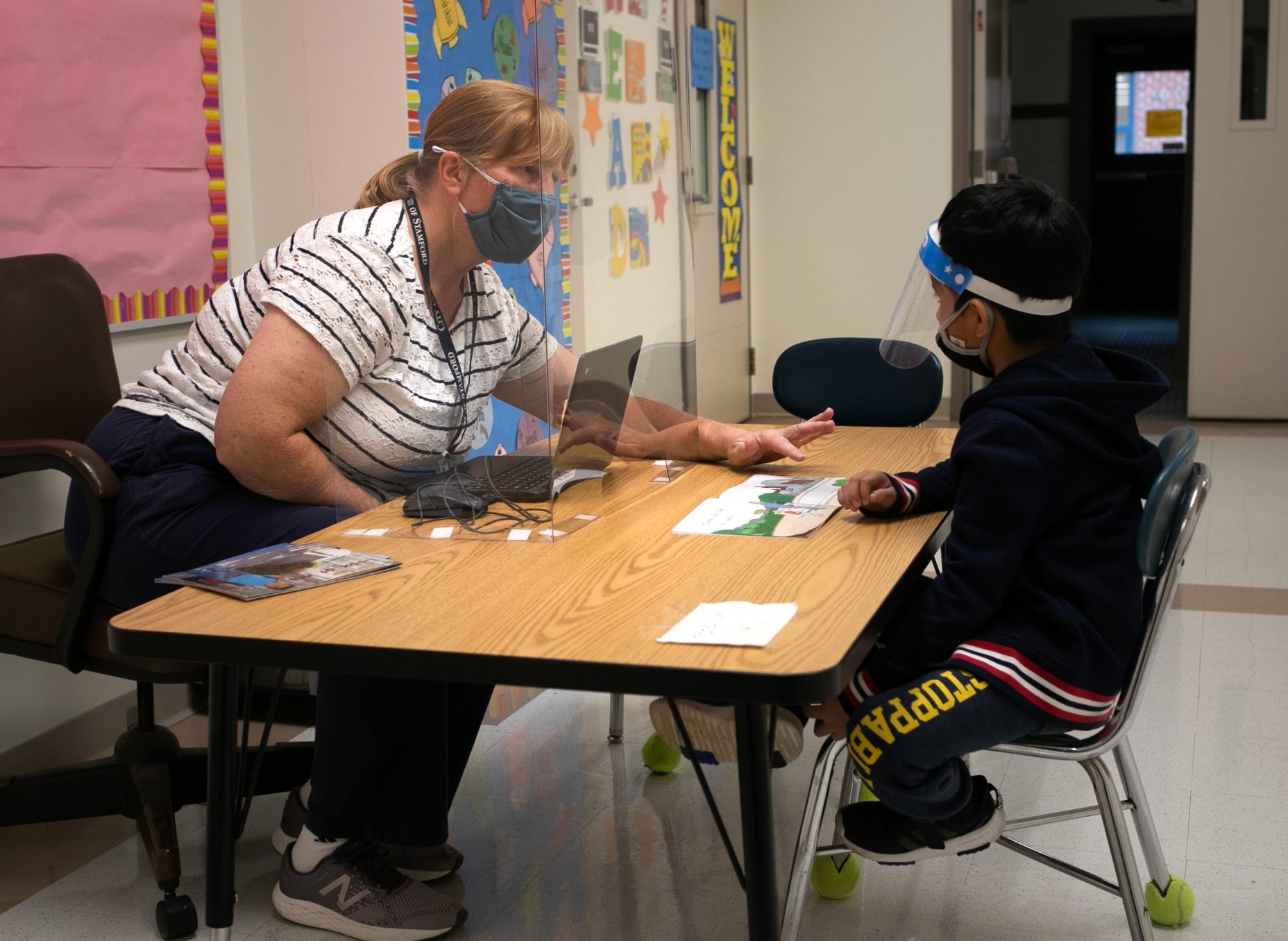 STAMFORD, CONNECTICUT - OCTOBER 21: Kindergarten teacher Maggie Peterson reaches around a safety shield while giving a reading test to a student at Stark Elementary School on October 21, 2020 in Stamford, Connecticut. Stamford Public Schools is continuing the fall semester with a hybrid model of in-class and distance learning, occasionally quarantining individual classes when a student or faculty member tests positive for COVID-19.  (Photo by John Moore/Getty Images)