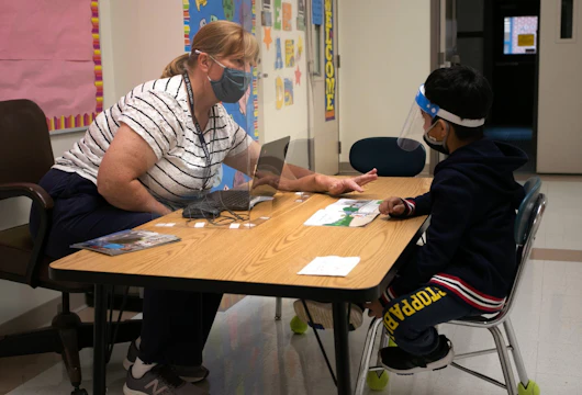 STAMFORD, CONNECTICUT - OCTOBER 21: Kindergarten teacher Maggie Peterson reaches around a safety shield while giving a reading test to a student at Stark Elementary School on October 21, 2020 in Stamford, Connecticut. Stamford Public Schools is continuing the fall semester with a hybrid model of in-class and distance learning, occasionally quarantining individual classes when a student or faculty member tests positive for COVID-19. (Photo by John Moore/Getty Images)