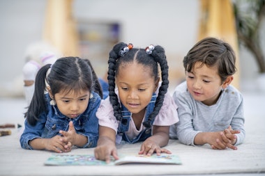 A group of three multi-ethnic preschool children lay on the floor intensely reading a story book together
