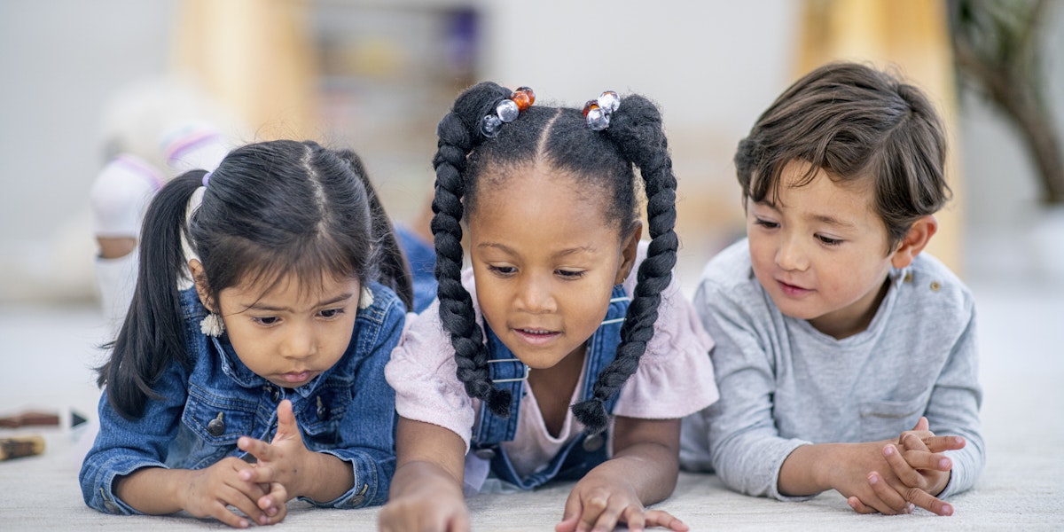 A group of three multi-ethnic preschool children lay on the floor intensely reading a story book together
