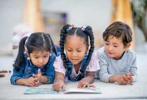 A group of three multi-ethnic preschool children lay on the floor intensely reading a story book together