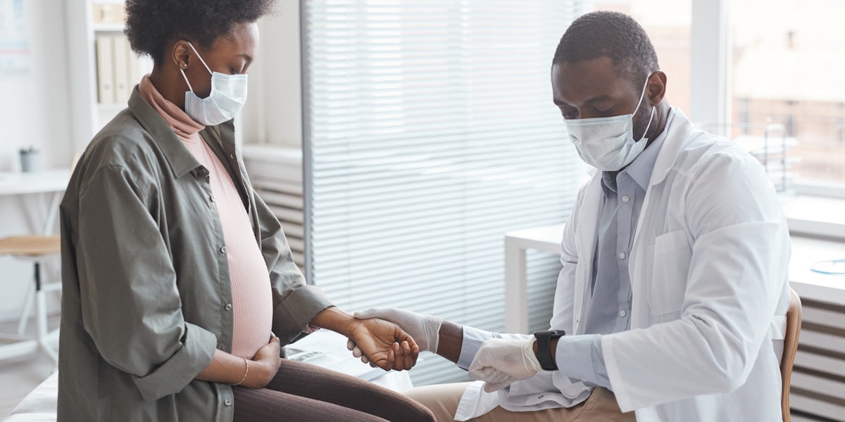 African young man in mask measuring the pulse of pregnant woman and caring about her health at hospital