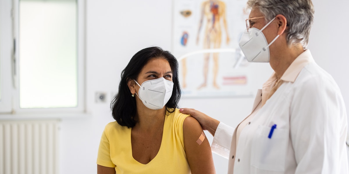 Mature woman visiting clinic for health checkup with a female doctor standing by. Senior doctor talking with female patient in clinic. Both wearing face masks.