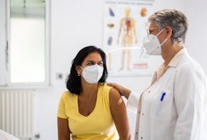 Mature woman visiting clinic for health checkup with a female doctor standing by. Senior doctor talking with female patient in clinic. Both wearing face masks.