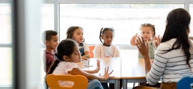 Sitting at a table in their classroom, a multi-ethnic group of preschool age children plays counting games with their unrecognizable female teacher.