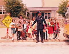 UNITED STATES - CIRCA 1960s:  Group of children at curb in front of school, waiting for signal from policeman.