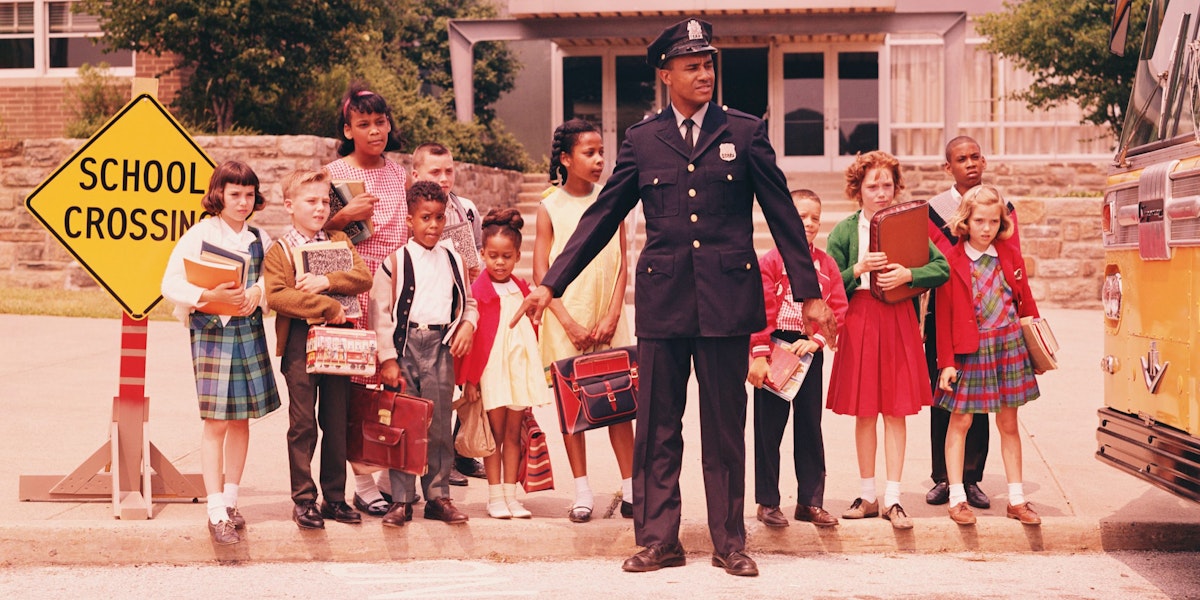 UNITED STATES - CIRCA 1960s:  Group of children at curb in front of school, waiting for signal from policeman.