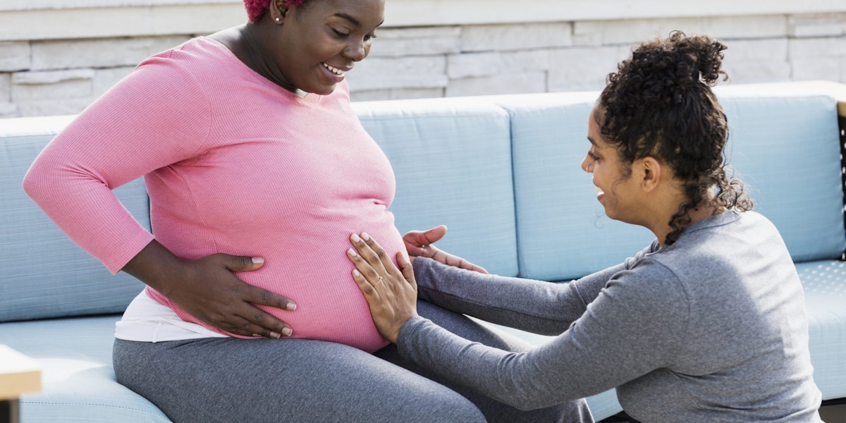 A pregnant African-American woman with her doula or birth support coach. The expectant mother, who had pink hair and is wearing a pink shirt, is sitting outdoors on a patio sofa smiling. The doula, a mixed race woman, is kneeling in front of her, touching her round abdomen. Both women are in their 30s.