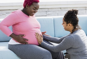 A pregnant African-American woman with her doula or birth support coach. The expectant mother, who had pink hair and is wearing a pink shirt, is sitting outdoors on a patio sofa smiling. The doula, a mixed race woman, is kneeling in front of her, touching her round abdomen. Both women are in their 30s.