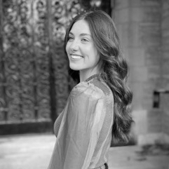 Adrianna, a white woman, stands outside, backgrounded by an iron gate and brick wall. She smiles and looks to the side of the camera. She has long auburn hair and wears a blue and purple shirt.
