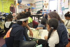 A teacher and two students are sitting on a classroom floor as the teacher holds a book for one of the students to read.