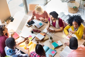 A group of students are sitting together at a wooden table talking and collaborating.
