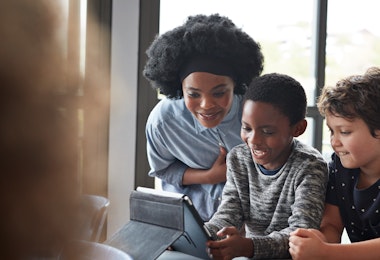 A Black teacher is looking at an iPad with two students.