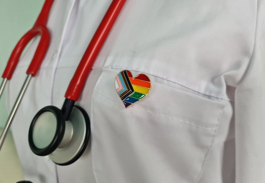 A close-up image of a doctor's coat with a heart-shaped pin that shows the Progress Pride Flag.