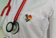 A close-up image of a doctor's coat with a heart-shaped pin that shows the Progress Pride Flag.