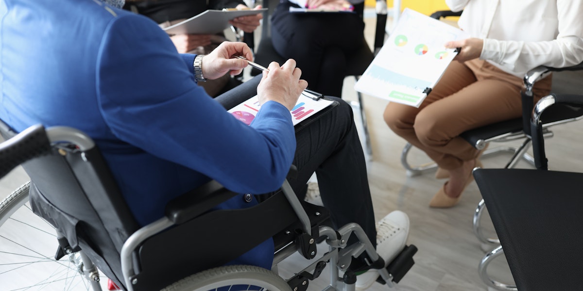 A close-up shot of a man in a wheelchair holding documents and speaking with coworkers.