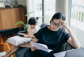 A young Asian mother looking stressed while managing her bills and documents, and her daughter is using a digital tablet in the background.