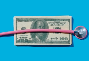 A pink stethoscope is on top of a pile of dollar bills against a blue background.