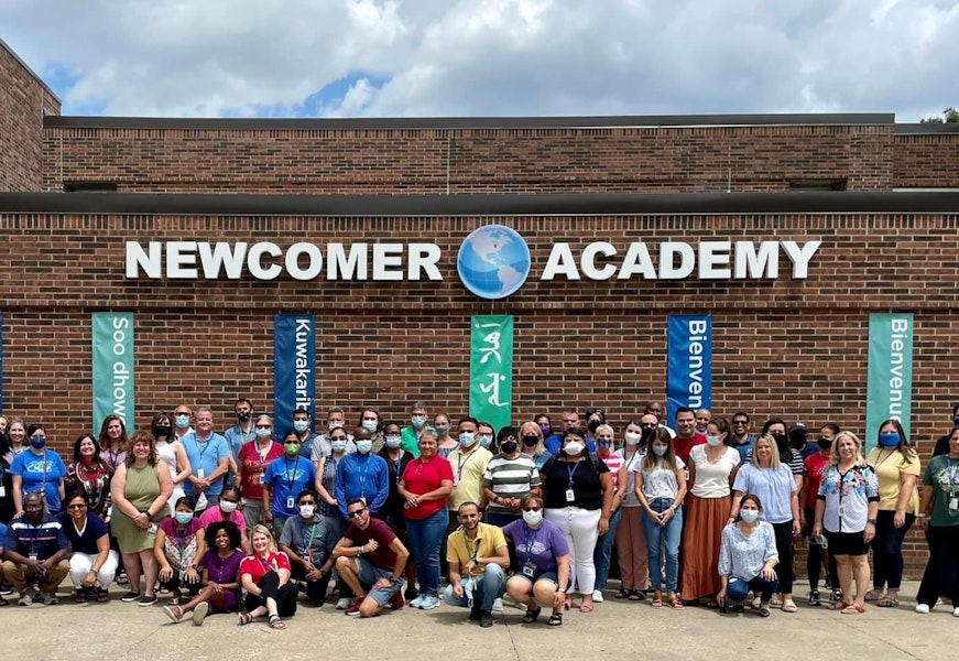 A group of teachers pose outside a school building that reads Newcomer Academy.