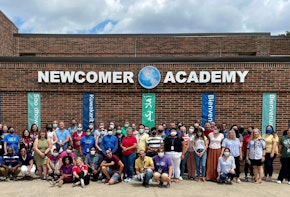 A group of teachers pose outside a school building that reads Newcomer Academy.