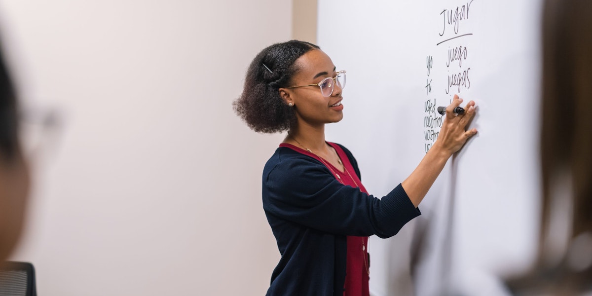 A young ethnic female professor stands at a large whiteboard and writes down Spanish verbs to conjugate.