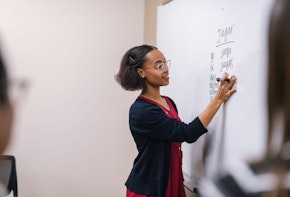 A young ethnic female professor stands at a large whiteboard and writes down Spanish verbs to conjugate.