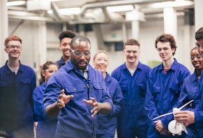 A tutor teaches his class about renewable energy in an engineering workshop. They are all wearing protective eyewear and blue coveralls.