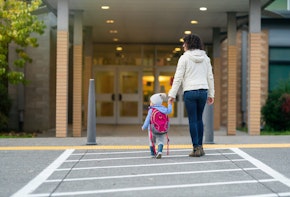 Rear view of a cheerful young mother holding her toddler daughter's hand while walking at crosswalk to the child's school.