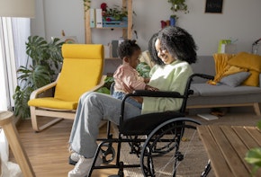 Toddler girl sitting on smiling mother with disability in wheelchair at home