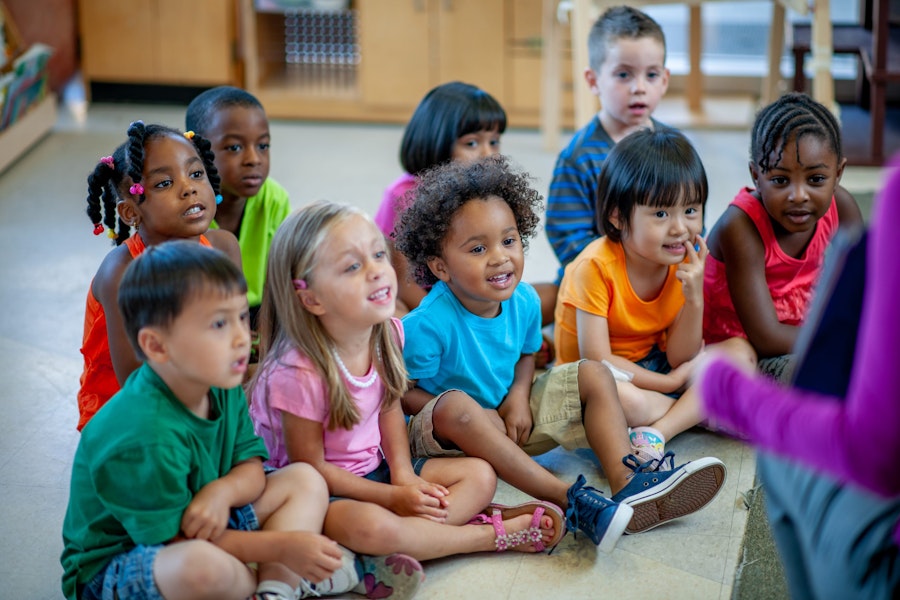 A daycare teacher sits with her students in front of her as she reads the group a story.  The children are each dressed casually in bright colours and focused on the story.