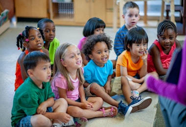 A daycare teacher sits with her students in front of her as she reads the group a story. The children are each dressed casually in bright colours and focused on the story.