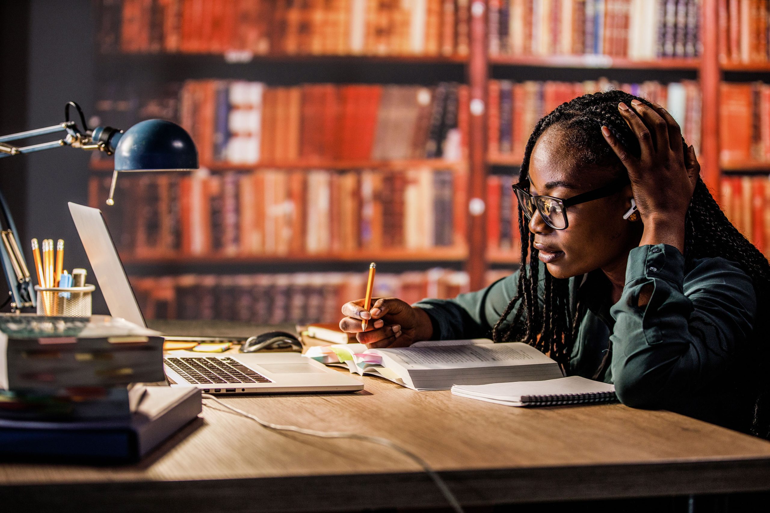 Copy space shot of annoyed young woman sitting at table in a library, head in hand, listening to music via blue-tooth headphones and studying for exam.