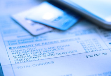 A hospital bill for $36,000 with a line item of various charges is photographed with a very shallow depth of field. A credit card and a mobile device rest out of focus in the background.