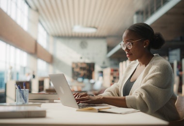 Dedicated African American Female Student Engaged in Online Learning at a Modern Library. Young Woman Using Laptop for Research, Surrounded by Books and Academic Environment.