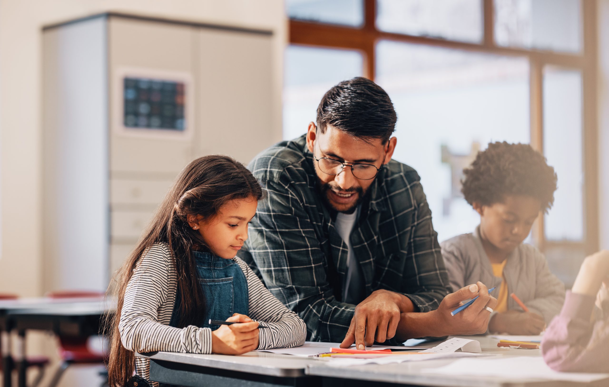 Man mentoring elementary school children. Teacher showing kids how to draw in a class. Male educator assists a young girl on how to complete an art task in a primary class.